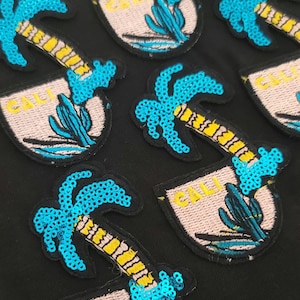 May include: Embroidered patches with a California theme. Each patch features a palm tree with blue sequins, a cactus, and the word "CALI" in yellow. The patches are on a black background.