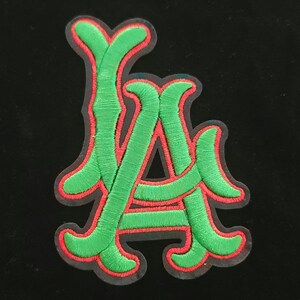 May include: Embroidered patch showing the letters "LA" in green with a red border. The patch has a detailed, textured appearance, and is set against a black background. This patch is suitable for jackets, bags, or other fabric items.
