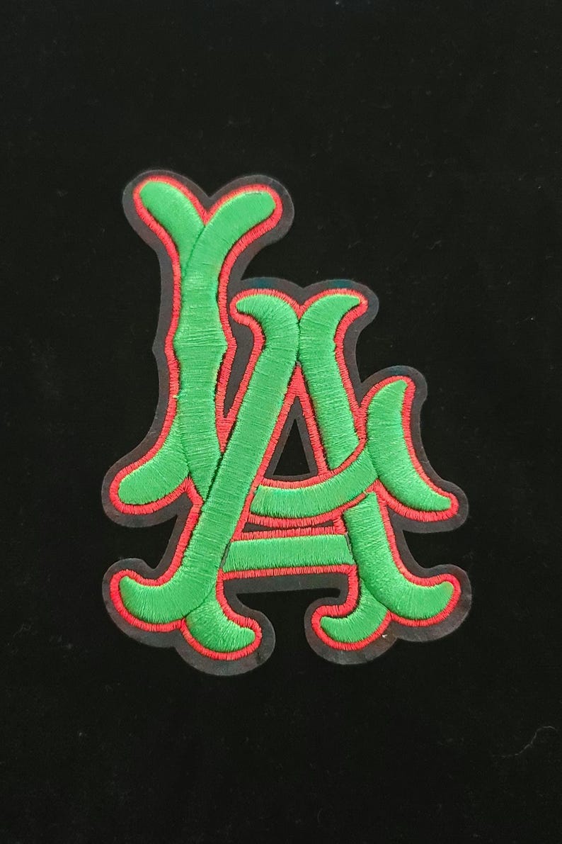 May include: Embroidered patch showing the letters "LA" in green with a red border. The patch has a detailed, textured appearance, and is set against a black background. This patch is suitable for jackets, bags, or other fabric items.