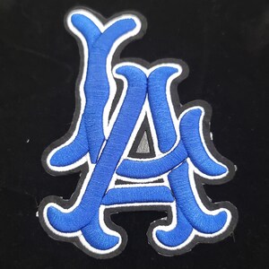 May include: An embroidered patch with the letters "LA" in blue, with a white border, on a black background. The letters are in a stylized script. This patch could be used on clothing or accessories.