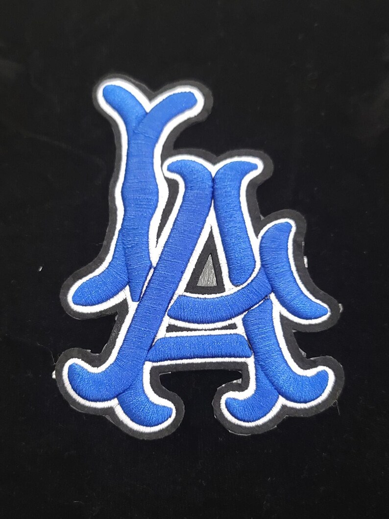 May include: An embroidered patch with the letters "LA" in blue, with a white border, on a black background. The letters are in a stylized script. This patch could be used on clothing or accessories.