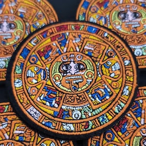 May include: Embroidered patch featuring a detailed Aztec calendar design. The patch is circular, with a black border and intricate patterns in shades of orange, yellow, blue, and green. The central figure has a face with a detailed design. The patches are likely for clothing or accessories.