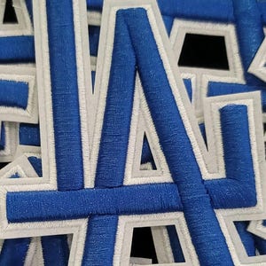 May include: Close-up of embroidered patches featuring the letters "LA" in blue with white outlining. The patches are densely packed, showcasing the detailed stitching and vibrant colors. The design is a classic sports team logo.