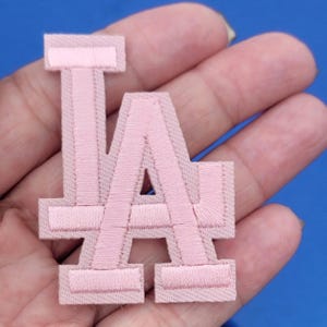 May include: A light pink embroidered patch displaying the letters "LA". The patch has a textured finish and a light pink border. It is designed for application on clothing or accessories, offering a decorative element.