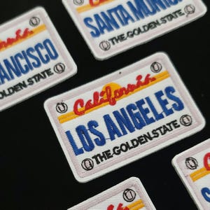 May include: Embroidered patches resembling California number plates. The patches display city names such as Los Angeles and Santa Monica, with the text "THE GOLDEN STATE". The patches have a white border.