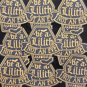 May include: A collection of dark blue embroidered patches with gold lettering. Each patch features the text "be a Lilith, not an Eve" in a gothic font, with bat and star accents. The patches are hexagonal with a banner design.