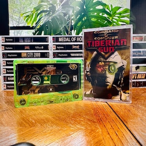 May include: A selection of retro video games, featuring a bright green cassette tape for "Tiberian Sun" and a game case with the same title. Other game cases are stacked in the background, with titles such as "Medal of Honour" and "Sim City 2000".