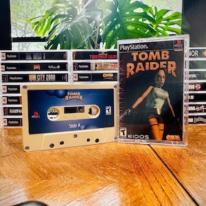 Puede incluir: Una colección de videojuegos retro y una cinta de cassette. El juego de PlayStation "Tomb Raider" se muestra de forma destacada, junto con otras carcasas de juegos. También se ve una cinta de cassette con la etiqueta "Tomb Raider". Los artículos están colocados sobre una superficie de madera.