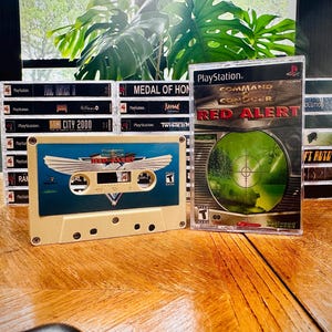 Op de afbeelding: Een verzameling vintage videogames, waaronder een PlayStation Command & Conquer: Red Alert game case en een cassettebandje. Het cassettebandje heeft een blauw en beige ontwerp met de titel van het spel. Andere game cases zijn zichtbaar.
