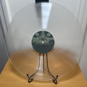 May include: A clear vinyl record on a black stand. The record has a green and black label with a creature on it.