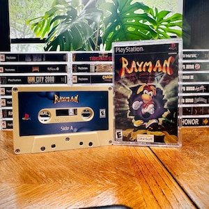 May include: A vintage Rayman cassette tape and a PlayStation game case are displayed on a wooden surface. The cassette tape is beige with a blue label, featuring the game's title. The game case shows Rayman bursting through a wall. Other game cases are in the background.