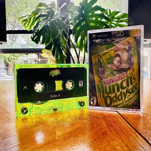 Puede incluir: Una cinta de cassette verde neón con la etiqueta "Side A" junto a una caja de juego de Xbox. La caja del juego presenta el título "Oddworld Munch's Oddysee" con un personaje de dibujos animados. Sobre una superficie de madera.