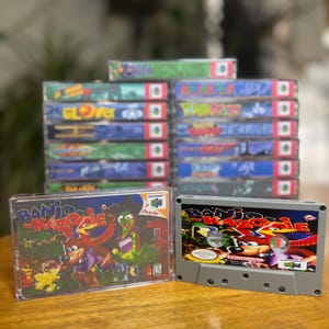 Puede incluir: Una pila de cartuchos de juegos de Nintendo 64, incluyendo Banjo-Kazooie, se muestra junto a una caja de plástico transparente con el cartucho de juego Banjo-Kazooie en su interior. También se muestra una cinta de cassette gris con la portada del juego Banjo-Kazooie.