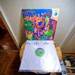 May include: A Nintendo 64 video game box for Banjo-Kazooie, featuring colorful cartoon characters and the game's title. A white record sleeve with a green and yellow label is in front of the game box.