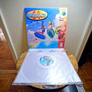 Puede incluir: Un disco de vinilo transparente con una etiqueta blanca que muestra el texto "Wave Race 64" en azul y blanco. El disco está empaquetado en una funda de plástico transparente con una caja de juego de Nintendo 64 que presenta un fondo azul y blanco con el texto "Wave Race 64" e imágenes de personajes montando motos acuáticas.