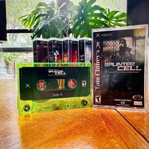 Op de afbeelding: Een neon-groen cassettebandje en een Xbox-gamecase voor "Tom Clancy's Splinter Cell" worden getoond. Op het cassettebandje staat "Side A" en de gamecase heeft een stealth-actie thema. Verschillende andere Xbox gamecases zijn zichtbaar op de achtergrond.