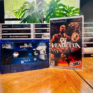 Può includere: Una musicassetta blu con l'etichetta "Def Jam Vendetta" accanto a una custodia di gioco per PlayStation 2 con lo stesso titolo. Dietro ci sono diverse altre custodie di giochi per PS2, con titoli come "God of War" visibili.
