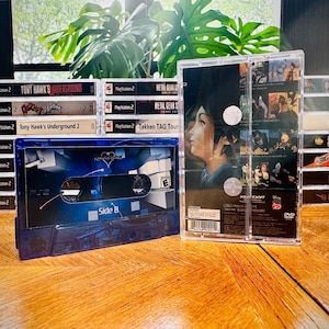 Peut inclure: Une collection de bo&icirc;tiers de jeux vid&eacute;o PlayStation 2 et une cassette bleue sur une surface en bois. Les jeux incluent "Tony Hawk's Underground" et "Metal Gear Solid". La cassette est &eacute;tiquet&eacute;e "Side B".