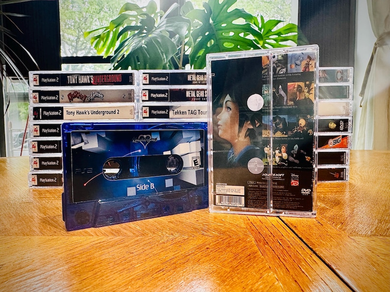 Peut inclure: Une collection de bo&icirc;tiers de jeux vid&eacute;o PlayStation 2 et une cassette bleue sur une surface en bois. Les jeux incluent "Tony Hawk's Underground" et "Metal Gear Solid". La cassette est &eacute;tiquet&eacute;e "Side B".