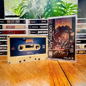 Puede incluir: Una colección de carcasas de videojuegos de PlayStation vintage y una cinta de cassette. La cinta de cassette presenta el juego "DOOM" con un diseño azul y beige. La caja del juego a la derecha también es "DOOM". Otros títulos de juegos son visibles.