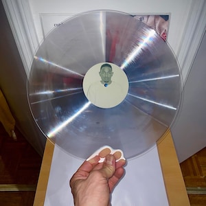 Puede incluir: Un disco de vinilo transparente con una etiqueta blanca que presenta un retrato de una persona con un fondo azul. El disco está en un estante de madera con otros discos.