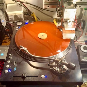 Puede incluir: Un tocadiscos negro Stanton ST.150 con un disco de vinilo naranja translúcido en el plato. Un conjunto de cabezal de corte metálico está posicionado sobre el disco, con varios cables electrónicos y componentes visibles en el fondo.