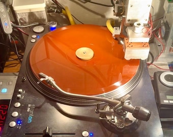 May include: A black Stanton ST.150 turntable with an orange vinyl record. The turntable has a silver tone arm and a control panel with buttons and a slider. A robotic arm is positioned above the record.