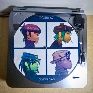 Peut inclure: Une platine vinyle argentée avec un disque vinyle noir et blanc représentant les membres du groupe Gorillaz de l'album "Demon Days".
