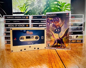 May include: A collection of retro video game media, including a PlayStation game case featuring a purple dragon and a cassette tape. The cassette tape has the text "Spyro" and "Side A". Other game cases are stacked in the background.