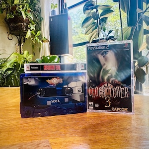 Może przedstawiać: Niebieska kaseta "Clock Tower" i pudełko z grą PlayStation 2 dla "Clock Tower 3" są wyświetlane. Na pudełku z grą widnieje twarz kobiety i logo Capcom. Widoczna jest również kaseta "Resident Evil".