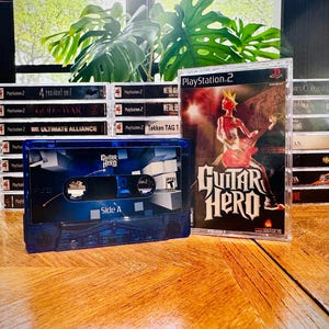 Peut inclure: Plusieurs jeux vidéo PlayStation 2, dont "Guitar Hero" et d'autres titres. Une cassette bleue avec l'inscription "Guitar Hero" est au premier plan. Les jeux sont empilés sur une surface en bois, avec une plante verte en arrière-plan.