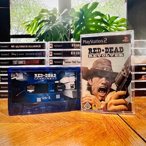 Könnte beinhalten: Eine Sammlung von PlayStation 2-Videospielen und einer Kassette. Die Spielhüllen sind gestapelt, mit Titeln wie "Ultimate Alliance" und "Tony Hawk's Underground". Die blaue Kassette zeigt den Spieltitel "Red Dead Revolver".