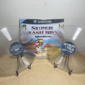 May include: Two clear vinyl records with a Super Smash Bros. Melee game case in the center. The records are on black stands.