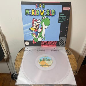 May include: A clear vinyl record with a label featuring a cartoon image of Mario and Yoshi from the Super Mario World video game. The label is surrounded by a white border with the text "Super Mario World" in black.