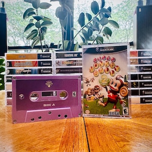Könnte beinhalten: Eine lila Kassette mit der Aufschrift "Seite A" neben einer Super Monkey Ball 2 Videospielhülle. Im Hintergrund sind mehrere andere GameCube-Spielhüllen gestapelt. Das Bild ist auf einer Holzoberfläche platziert.