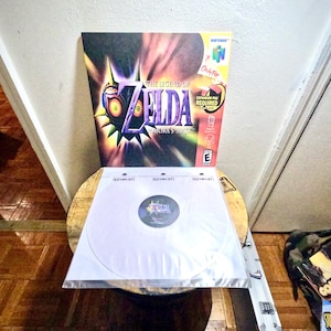 May include: A white vinyl record with a purple and black design in the center. The record is on a wooden table with a Nintendo 64 game cartridge for The Legend of Zelda: Majora's Mask in the background.