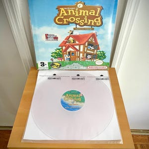 Może przedstawiać: Obudowa gry wideo Nintendo GameCube dla "Animal Crossing" jest wyświetlana na drewnianej półce. Obudowa gry zawiera kolorowe grafiki domu i postaci. Poniżej znajduje się przezroczysta płyta winylowa z etykietą "Animal Crossing".