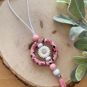 May include: A pink and floral patterned keychain with a white daisy in the center. The keychain has a pink tassel, white and pink beads, and a white cord with a pink magnetic clasp.
