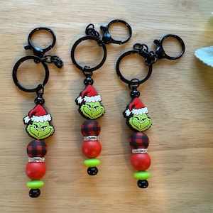 May include: Three keychains featuring a green Grinch face wearing a Santa hat. Each keychain has a black ring and clasp, with red and black plaid beads, red beads, and green beads. The keychains are on a wooden surface.