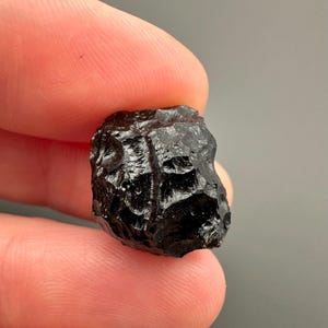 May include: A small, dark, irregularly shaped mineral specimen. The stone is a deep black colour with a glossy, reflective surface. The texture appears rough and uneven, with visible indentations and ridges. The mineral is held between two fingers against a grey background.