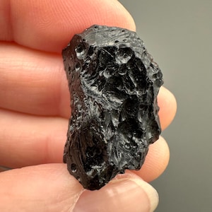 May include: A dark, irregularly shaped, black stone with a rough, textured surface. The stone has a glossy appearance and is held in a person's hand. The stone's surface has small, crater-like indentations.