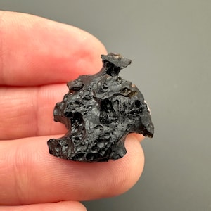 May include: A black, irregularly shaped piece of tektite with a rough, textured surface. The tektite has a pointed top and a rounded base.