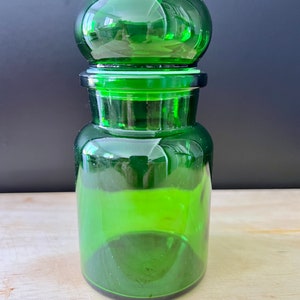 Puede incluir: Una botella de vidrio verde con un tapón redondo de vidrio verde. La botella es transparente y tiene una superficie texturizada.