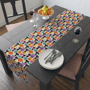 May include: A Halloween-themed table runner featuring a repeating pattern of black cats, jack-o'-lanterns, and colorful trees on a white background. The runner is displayed on a dark wooden table, with plates, silverware, and wine glasses.