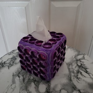 May include: Purple and pink tissue box cover with a textured, bubble-like design. The cover is made of fabric and has a square shape.
