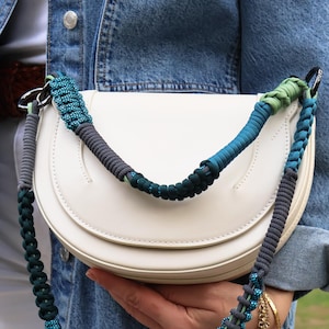 Puede incluir: Bolso de mano blanco semicircular con una correa trenzada colorida. La correa presenta tonos de verde azulado, verde y gris. El bolso se sostiene contra una chaqueta vaquera, lo que sugiere un accesorio informal.
