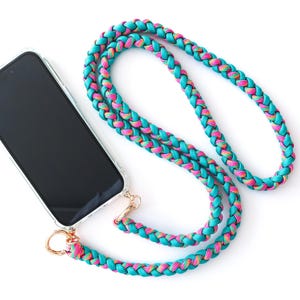 Op de afbeelding: Een doorzichtige telefoonhoes met een zwart scherm is bevestigd aan een turquoise, roze en gele gevlochten lanyard. De lanyard heeft een goudkleurige sluiting en ring. De telefoon en lanyard liggen op een witte achtergrond.