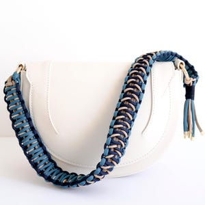 May include: A white, semi-circular handbag with a braided strap in shades of blue and beige. The strap is attached to the bag with gold-coloured hardware. The strap ends in a tassel with gold accents. The bag is made of a smooth material.