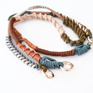 May include: Three macrame bag straps in shades of brown, beige, and teal, with gold-coloured metal clasps. The straps are made of tightly woven cord, showcasing a variety of knotting patterns. The background is plain white.