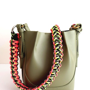 May include: An olive green bucket bag featuring a thick, braided shoulder strap made of neon pink, bright yellow, and black cord. The bag has a smooth, matte finish with structured side panels and metallic hardware rings connecting the strap.